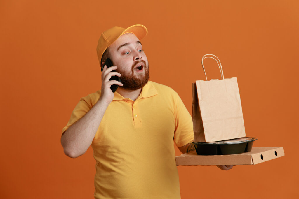 entregador de delivery em fundo amarelo segurando um pacote de delivery e atendendo um telefone.