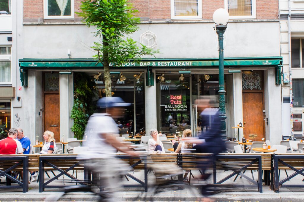 Fachada de restaurante em avenida movimentada, com várias pessoas passando de bicicleta e a pé na frente. 