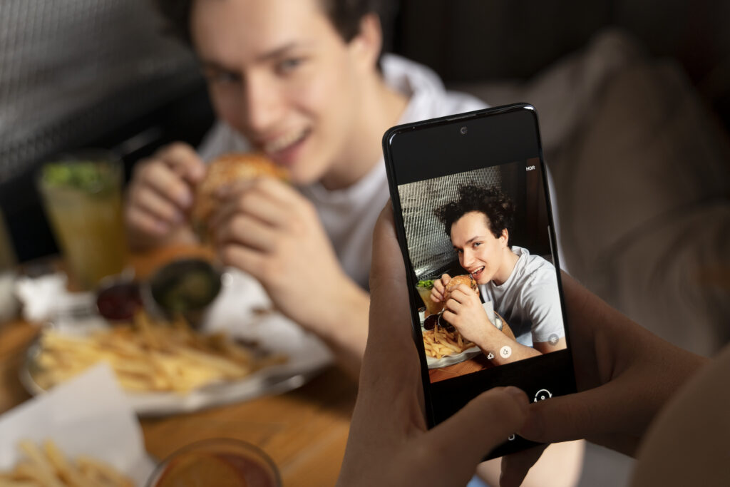 Celular tirando foto de homem mordendo um hamburguer.