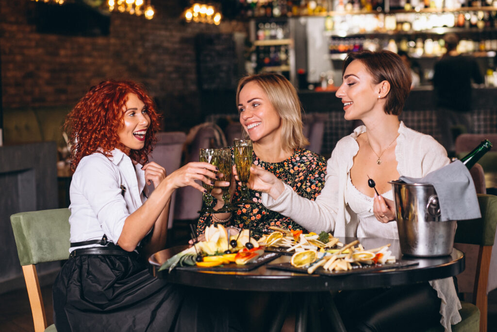 Três mulheres em restaurante, brindando taças de vinho e porções de degustação na mesa.