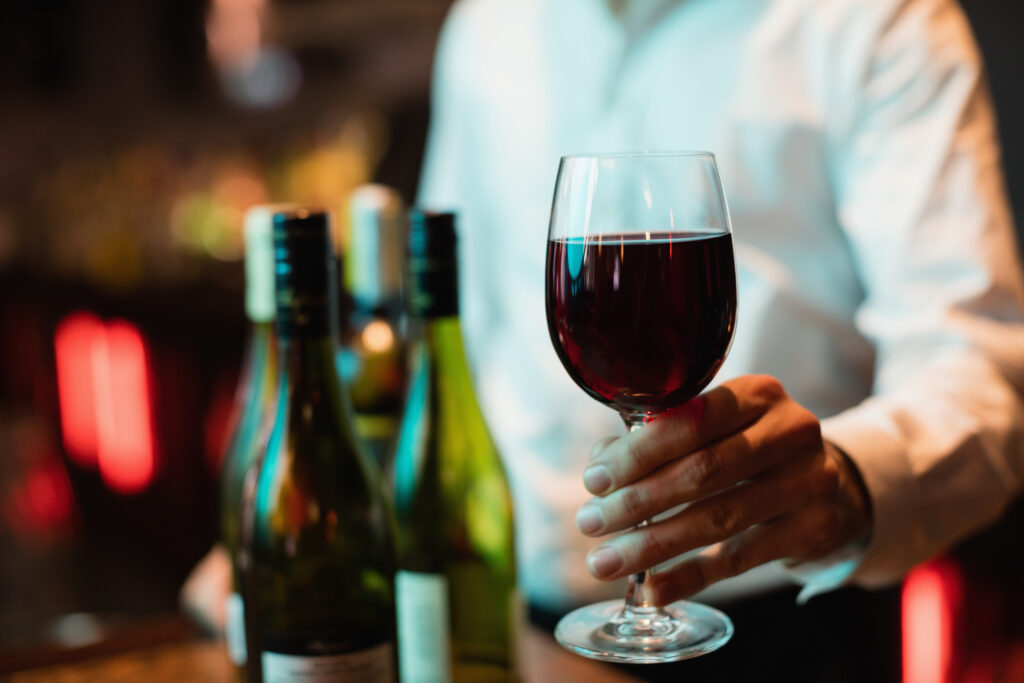 Close em mão segurando taça de vinho, ao fundo restaurante e uma mesa com três garrafas diferentes.