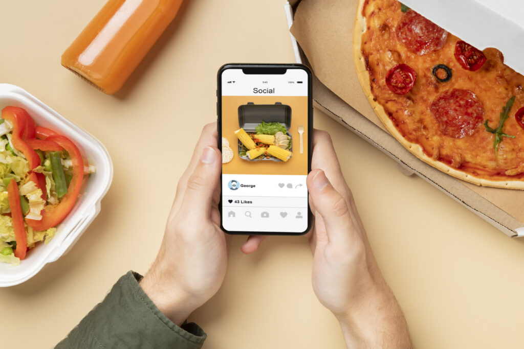 mãos segurando celular em cima de uma mesa com pizza de delivery, suco e salada em embalagem de entrega; 