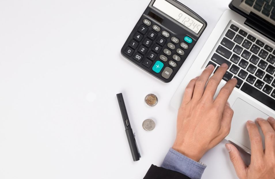 Foto tirada de cima; uma mesa com calculadora, caneta, moedas e notebook em cima.