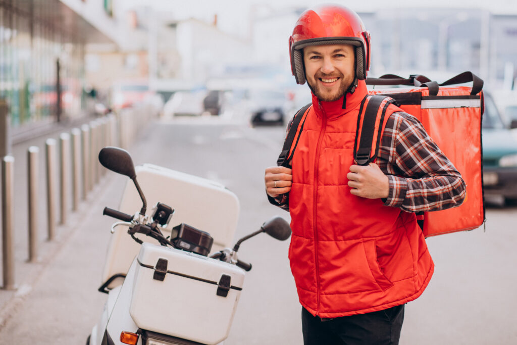 Motoboy com bolsa de entrega e moto estacionada, sorrindo para a câmera.
