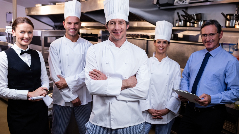 Chef, cozinheiros e equipe de atendimento sorrindo para foto na cozinha de restaurante.