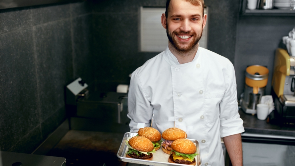 Chef segurando bandeja de hambúrgueres e sorrindo para foto. 