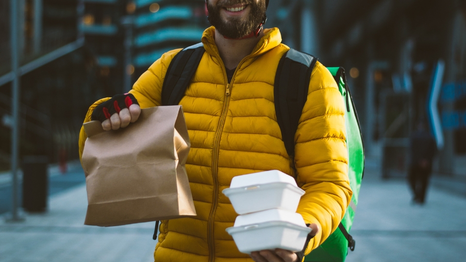 Entregador delivery na calçada sorrindo para foto com embalagem de comida em mãos.