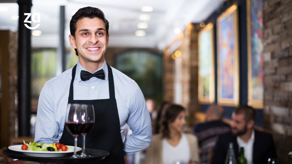 Garçom de social, gravata e avental segurando bandeja com duas taças de vinho e prato de salada em restaurante.