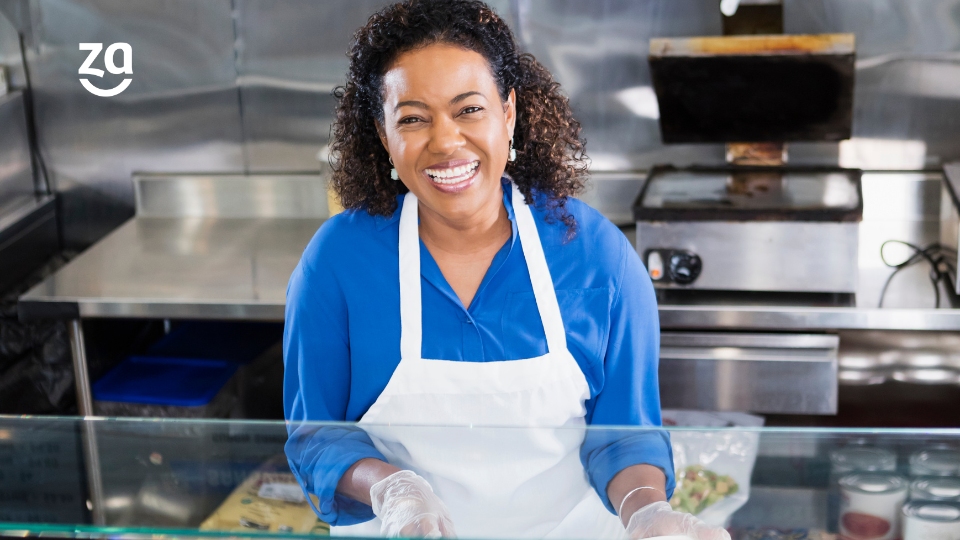 Empreendedora de avental sorrindo intensamente para foto, de avental na cozinha de seu restaurante.
