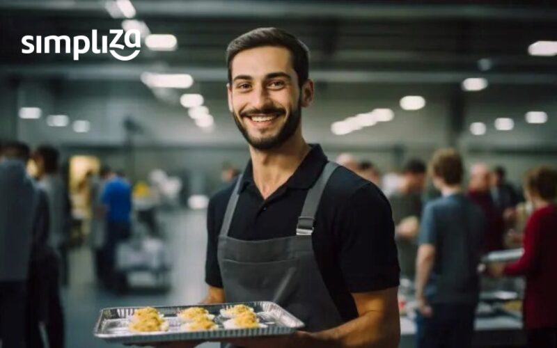 Foto de funcionário de empresa com uma bandeja na mão sorrindo, atrás dele está o refeitório de uma indústria com uma estação de alimentação para refeições coletivas atrás.