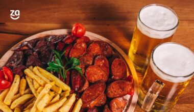 porção de batata com calabresa, tomate cereja e salsinha;porção em mesa de madeira e ao lado duas cervejas