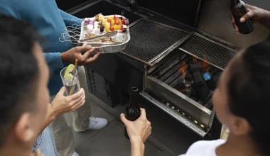 algumas pesoas em volta de uma churrasqueira com copos de bebida nas mãos. Uma pessoa está segurando um prato de comida