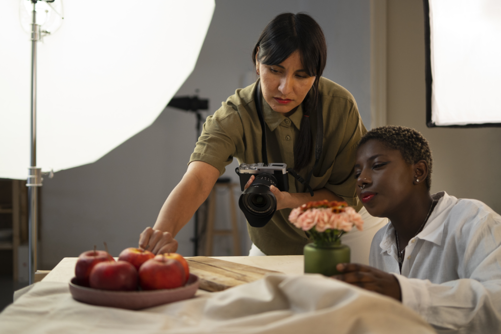 uma pessoa com uma camera na mão arrumando frutas na mesa para tirar foto, enquanto outra pessoa esta sentada na mesa olhando