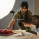 uma pessoa com uma camera na mão arrumando frutas na mesa para tirar foto, enquanto outra pessoa esta sentada na mesa olhando